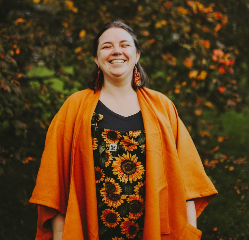 Woman wearing an orange cardigan over a sunflower-patterned dress with a blurred green and orange foliage background.